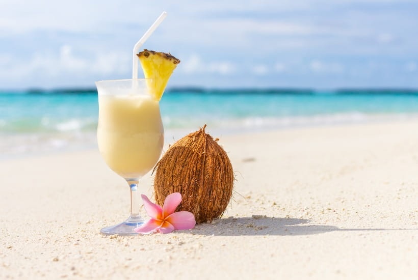 szklanka z drinkiem, obok szklanki lezy kokos, drink ozdobiony kawałkiem ananasa. Wszystko w scenerii nadmorskiej- na plaży.